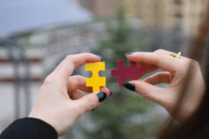 Hands holding yellow and maroon puzzle pieces, representing the hidden truths of grief, the pieces we carry alone, and the slow process of finding what fits.